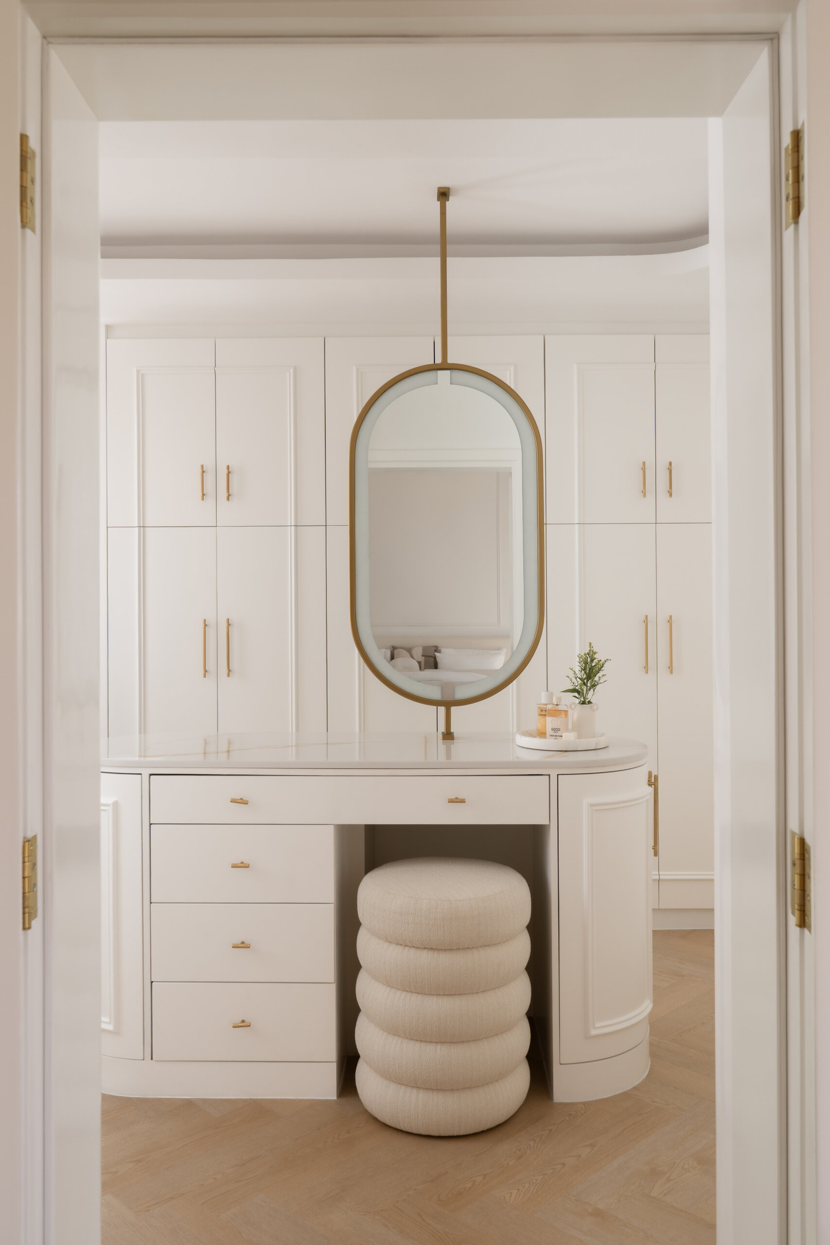 A luxurious walk-in closet and dressing room inside The White Arcade in Dubai. Designed by D’Ora Tokai Designs, this elegant space features custom white wardrobes, curved cabinets, brass hardware, a bespoke vanity table, oval mirror, and sculptural upholstered stool.