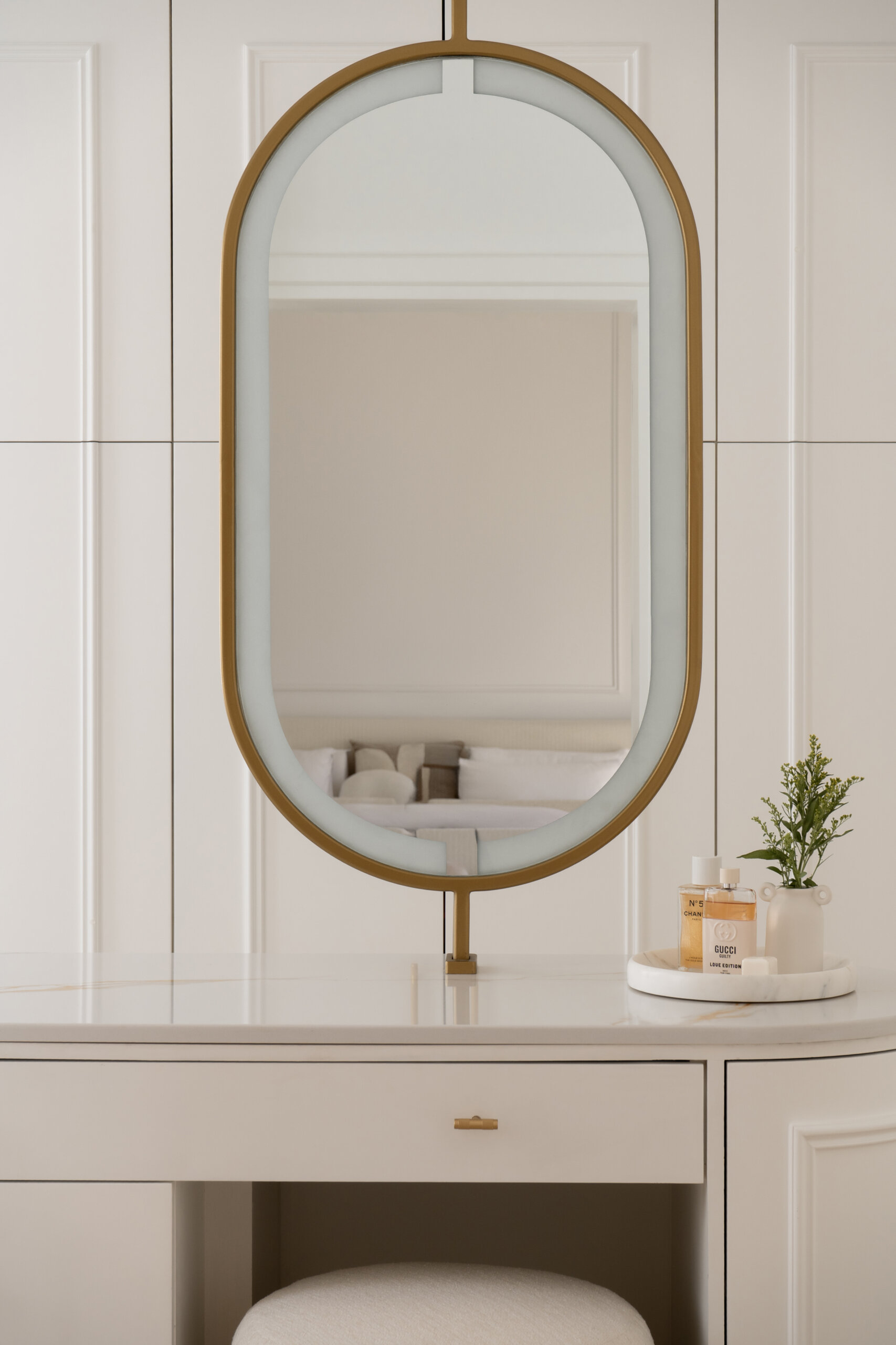 A luxurious walk-in closet and dressing room inside The White Arcade in Dubai. Designed by D’Ora Tokai Designs, this elegant space features custom white wardrobes, curved cabinets, brass hardware, a bespoke vanity table, oval mirror, and sculptural upholstered stool.