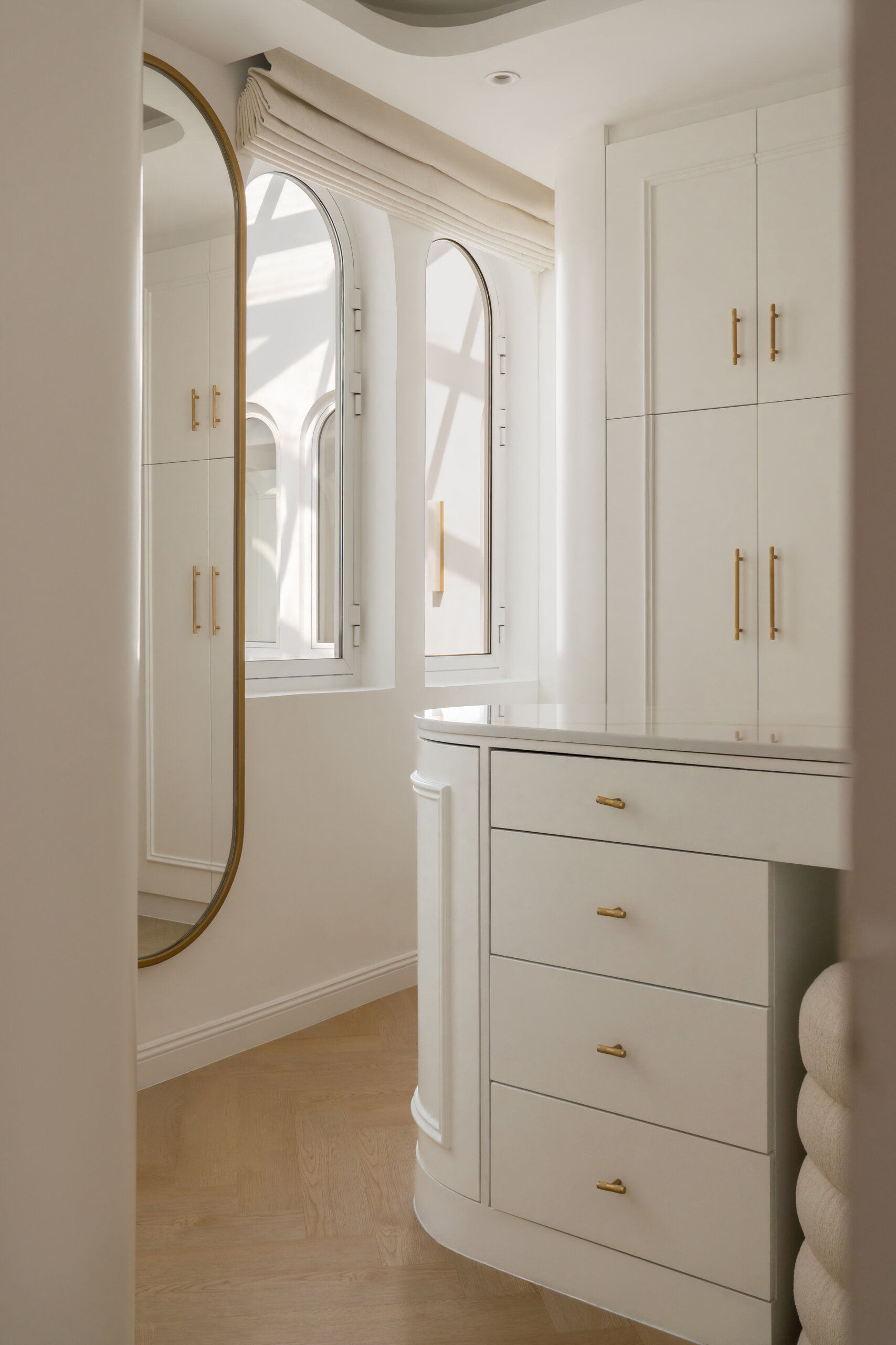 A luxurious walk-in closet and dressing room inside The White Arcade in Dubai. Designed by D’Ora Tokai Designs, this elegant space features custom white wardrobes, curved cabinets, brass hardware, a bespoke vanity table, oval mirror, and sculptural upholstered stool.
