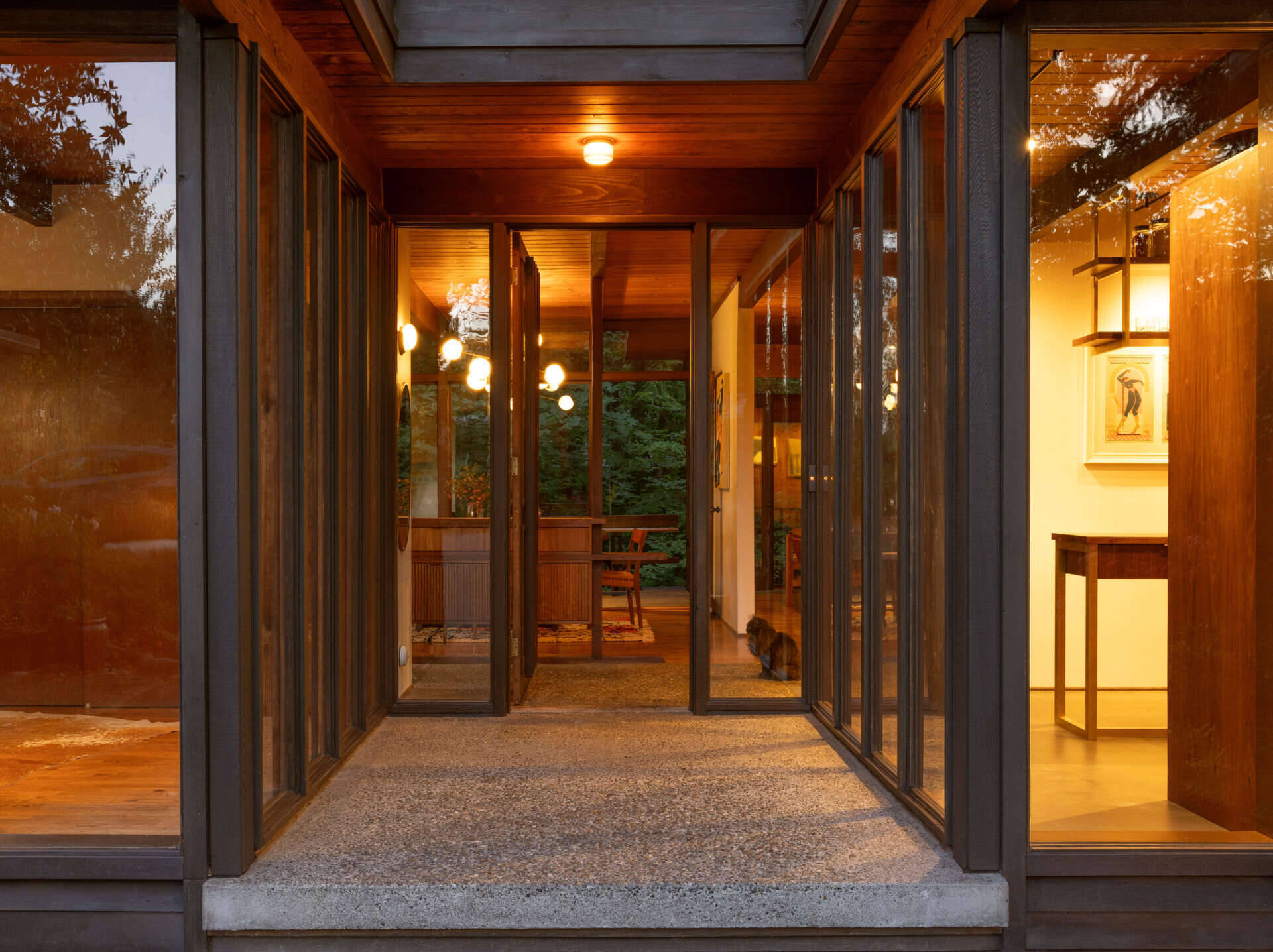 Step inside The Burrows, a renovated mid-century home in Seattle by chadbourne + doss architects. A recessed entrance features floor-to-ceiling glass, exposed timber ceilings, a concrete landing, and a custom walnut room divider.