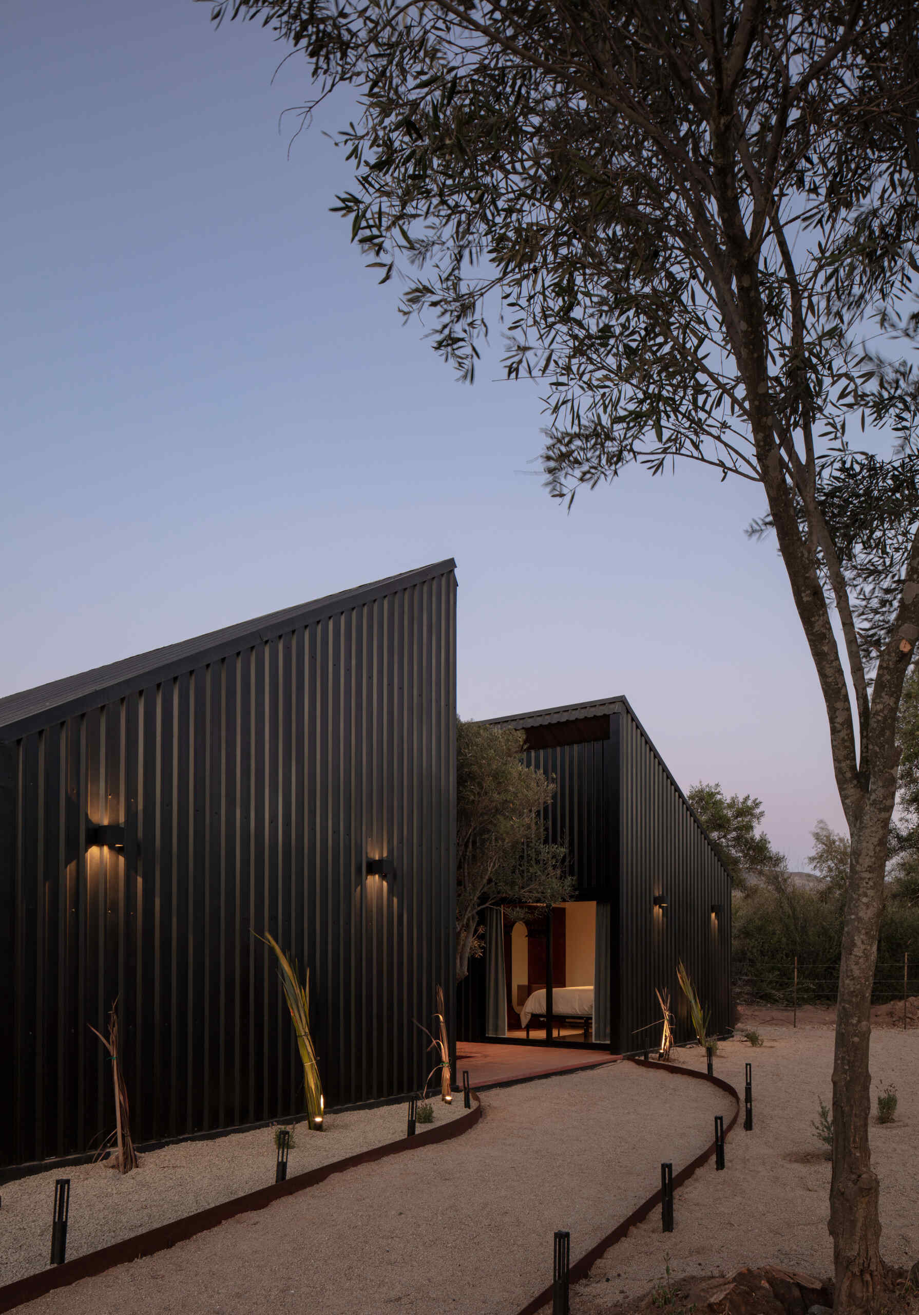 Explore the minimalist architecture of Serentha Cabin by ORTO STUDIO in Baja California, Mexico. Dark corrugated metal exteriors, sloping roofs, warm exterior lighting, gravel pathways, and olive trees create a modern rural cabin with an industrial edge.