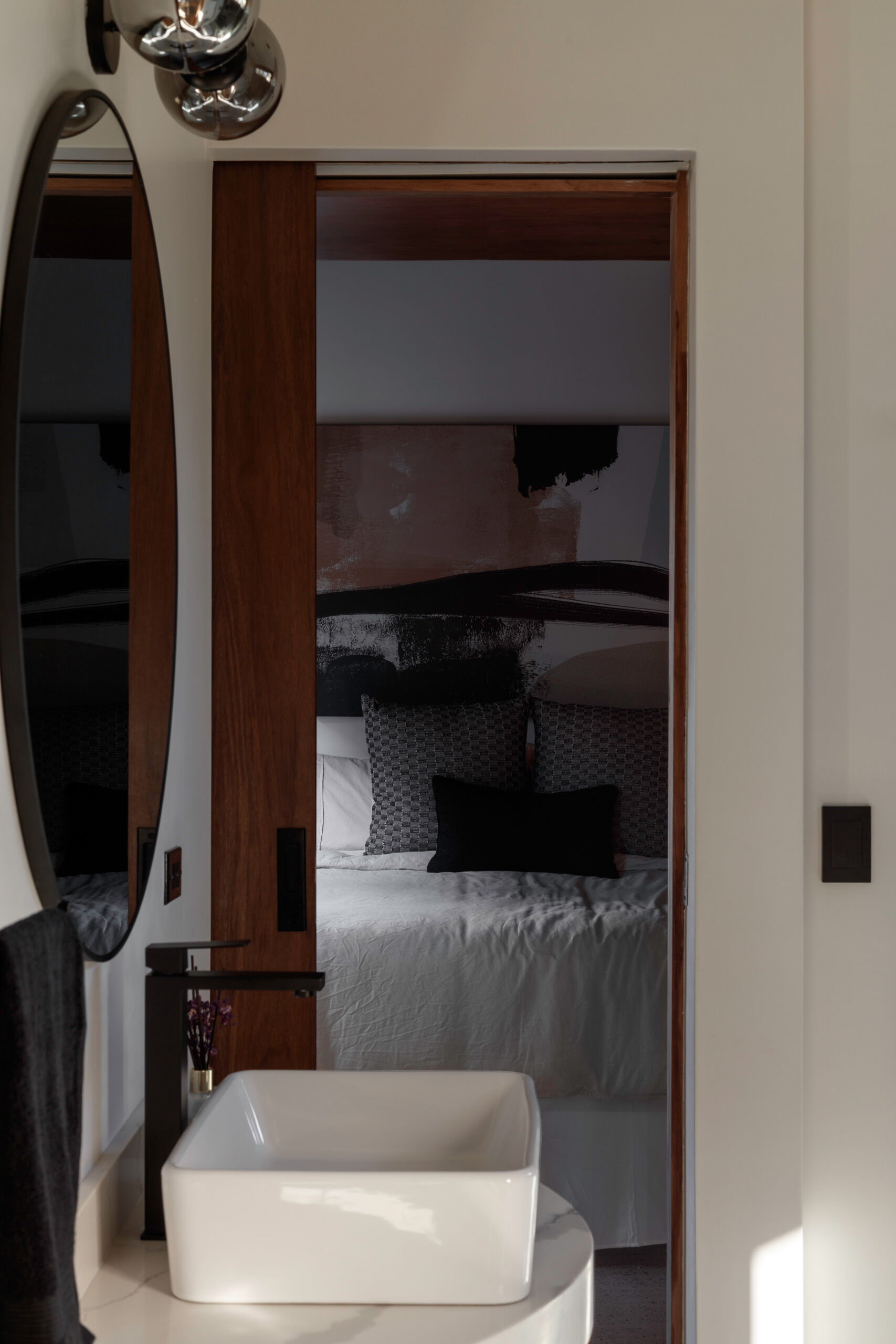 Explore the bedroom and bathroom at Serentha Cabin by ORTO STUDIO in Baja California, Mexico. Neutral bedding, abstract wall art, a wooden sliding door, an oval bathroom mirror, and a white vessel sink create a cozy, contemporary interior.