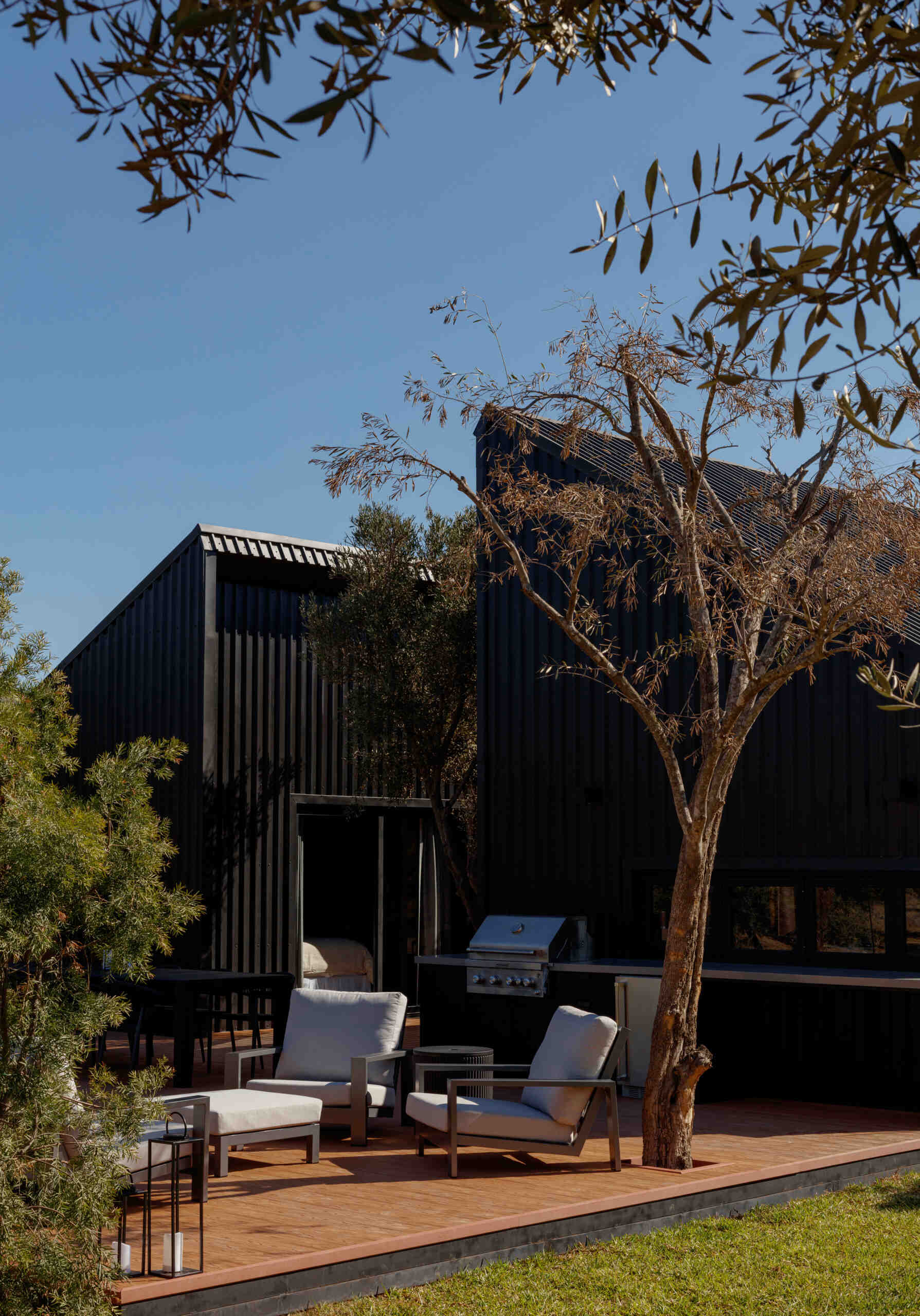 Discover the outdoor deck and fire pit at Serentha Cabin by ORTO STUDIO in Baja California, Mexico. Timber decking, black Adirondack chairs, a circular fire pit, and comfortable outdoor lounge furniture create an inviting space for relaxing and entertaining among the olive trees.