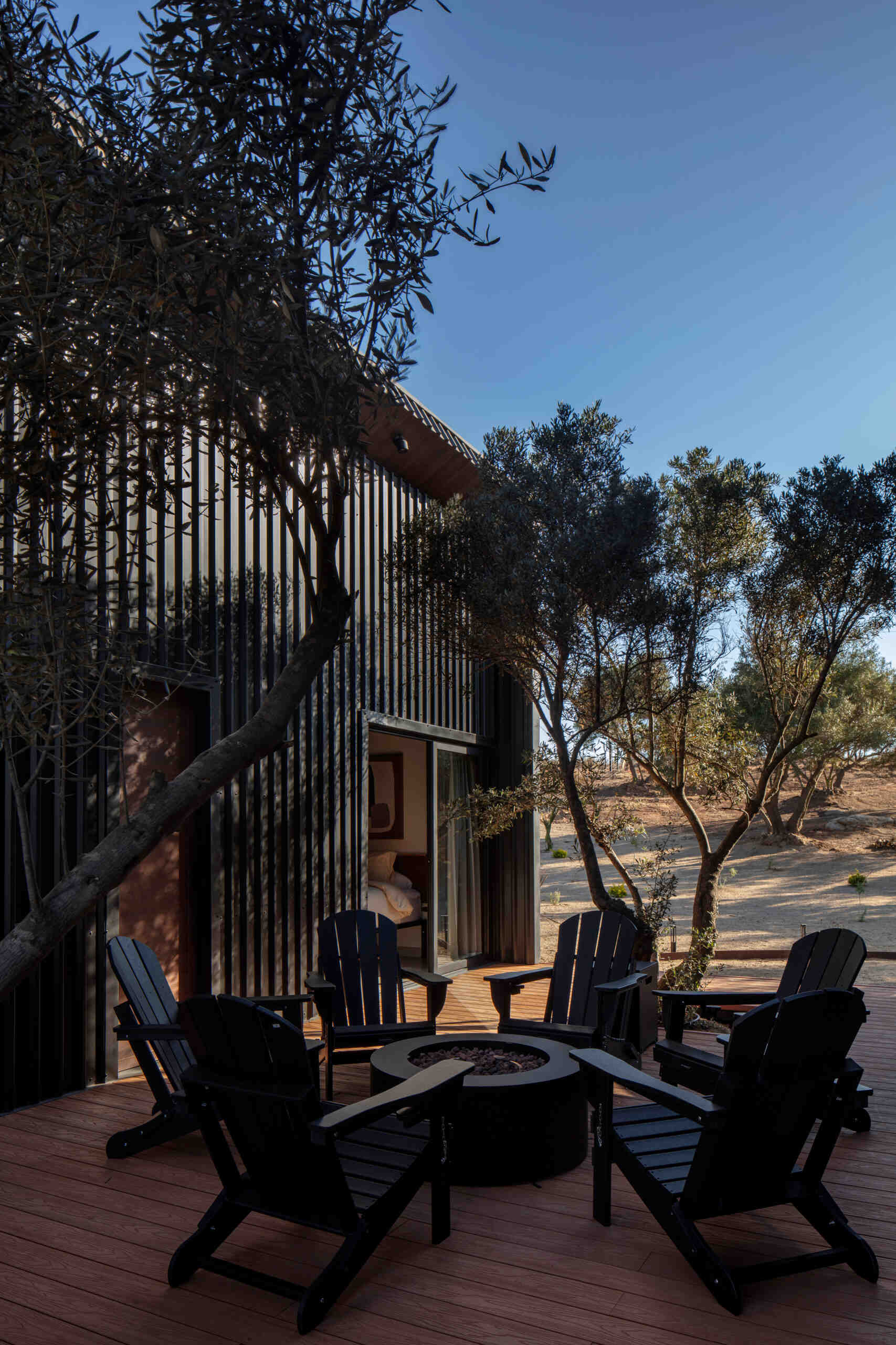 Discover the outdoor deck and fire pit at Serentha Cabin by ORTO STUDIO in Baja California, Mexico. Timber decking, black Adirondack chairs, a circular fire pit, and comfortable outdoor lounge furniture create an inviting space for relaxing and entertaining among the olive trees.