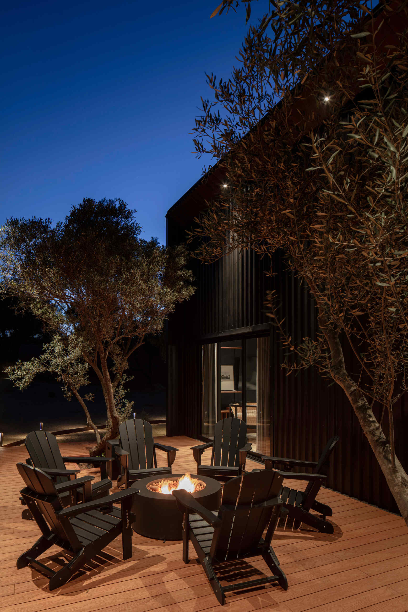 Discover the outdoor deck and fire pit at Serentha Cabin by ORTO STUDIO in Baja California, Mexico. Timber decking, black Adirondack chairs, a circular fire pit, and comfortable outdoor lounge furniture create an inviting space for relaxing and entertaining among the olive trees.