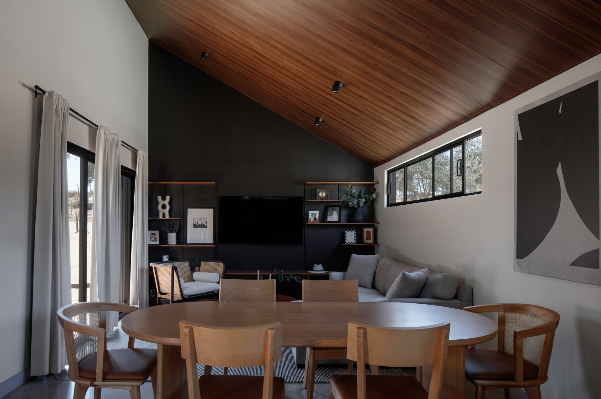 A modern dining area inside Serentha Cabin by ORTO STUDIO in Baja California, Mexico. An oval wooden dining table, six mixed timber and cane-back chairs, and oversized abstract wall art create a relaxed dining space within the cabin's open-plan interior.