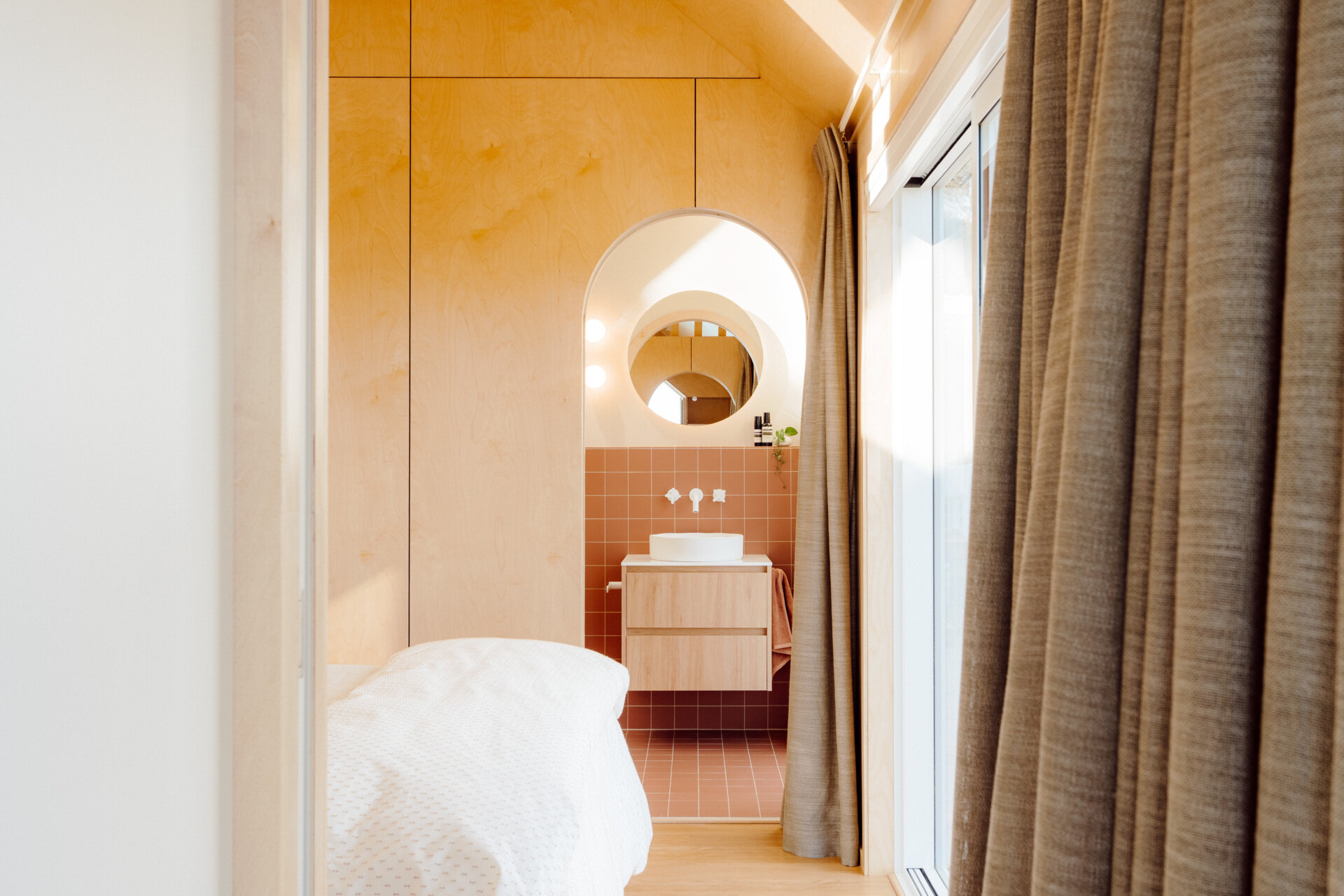 Discover a modern ensuite bathroom featuring terracotta square tiles, an arched doorway, a floating timber vanity, circular illuminated mirror and skylit walk-in shower. Small bathroom inspiration with warm colours and contemporary finishes.