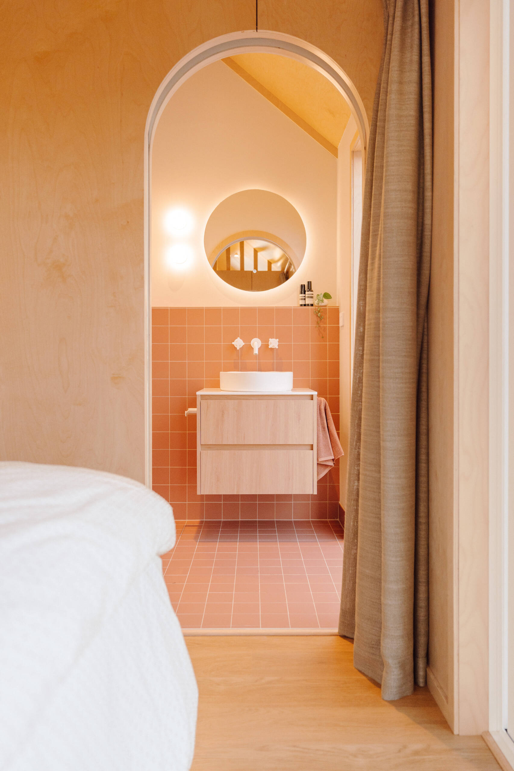 Discover a modern ensuite bathroom featuring terracotta square tiles, an arched doorway, a floating timber vanity, circular illuminated mirror and skylit walk-in shower. Small bathroom inspiration with warm colours and contemporary finishes.