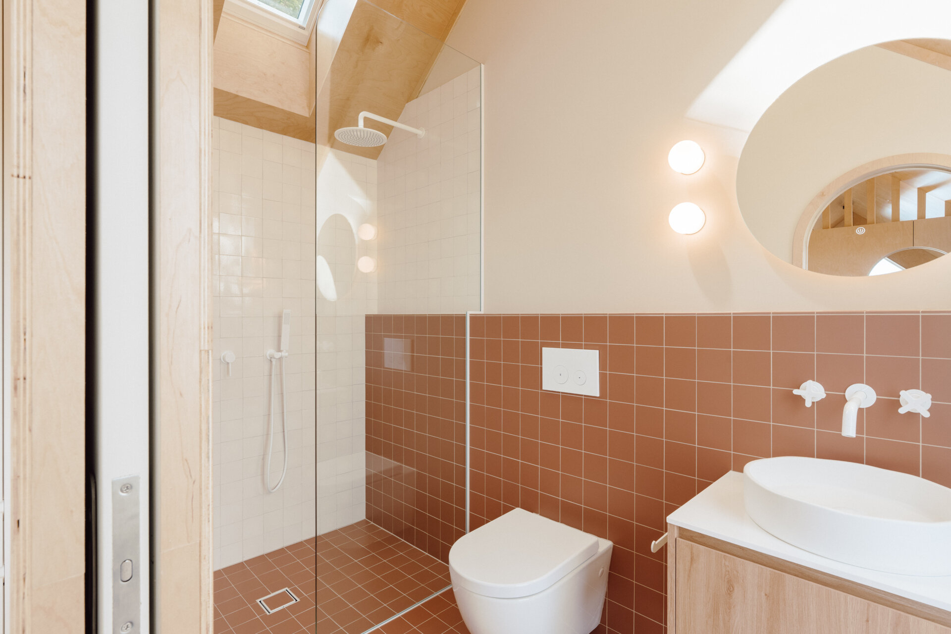 Discover a modern ensuite bathroom featuring terracotta square tiles, an arched doorway, a floating timber vanity, circular illuminated mirror and skylit walk-in shower. Small bathroom inspiration with warm colours and contemporary finishes.