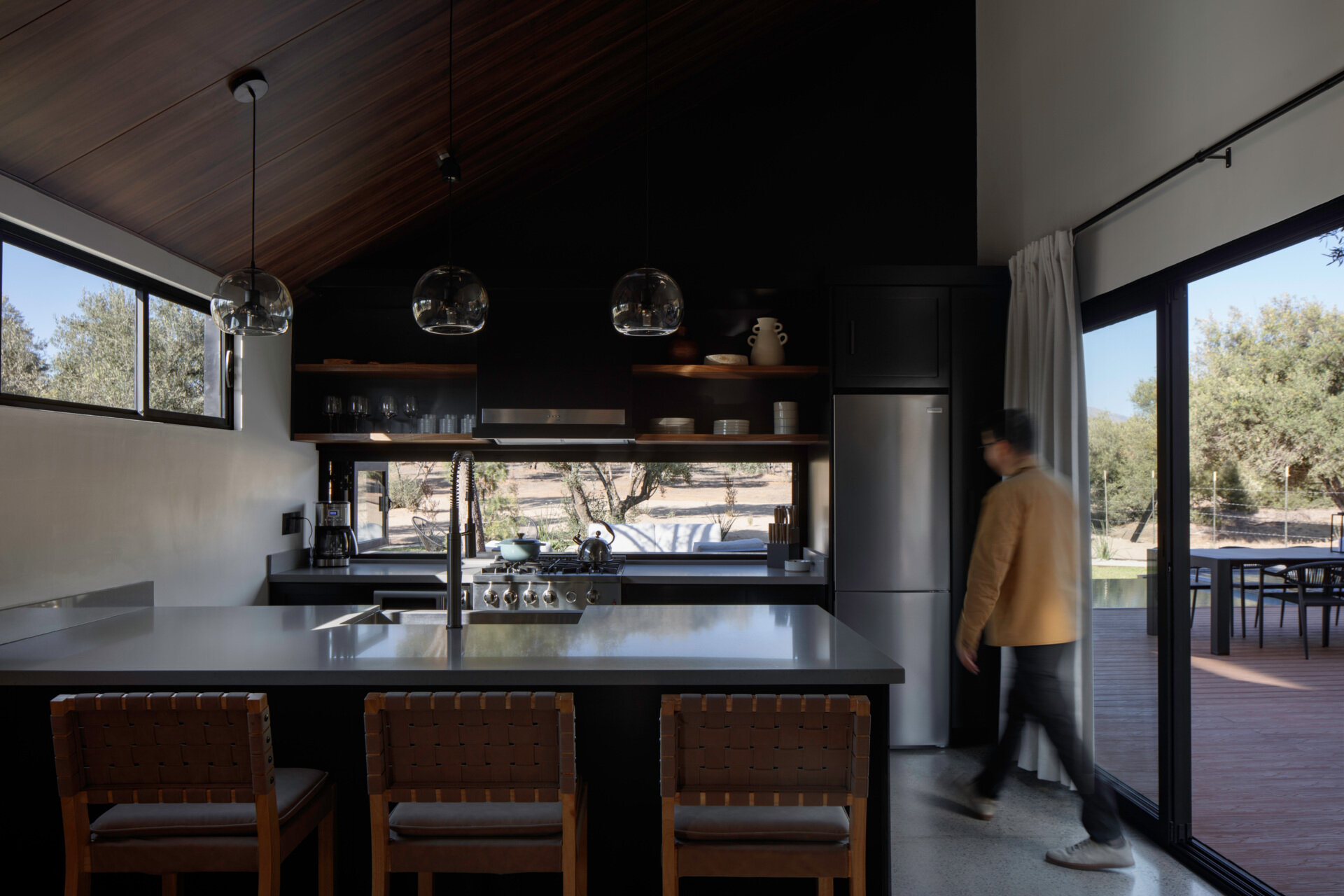 Explore the modern black kitchen at Serentha Cabin by ORTO STUDIO in Baja California, Mexico. Matte black cabinets, a large gray kitchen island, leather-woven bar stools, glass pendant lights, stainless steel appliances, and open timber shelves create a stylish cabin kitchen with views of the olive grove.