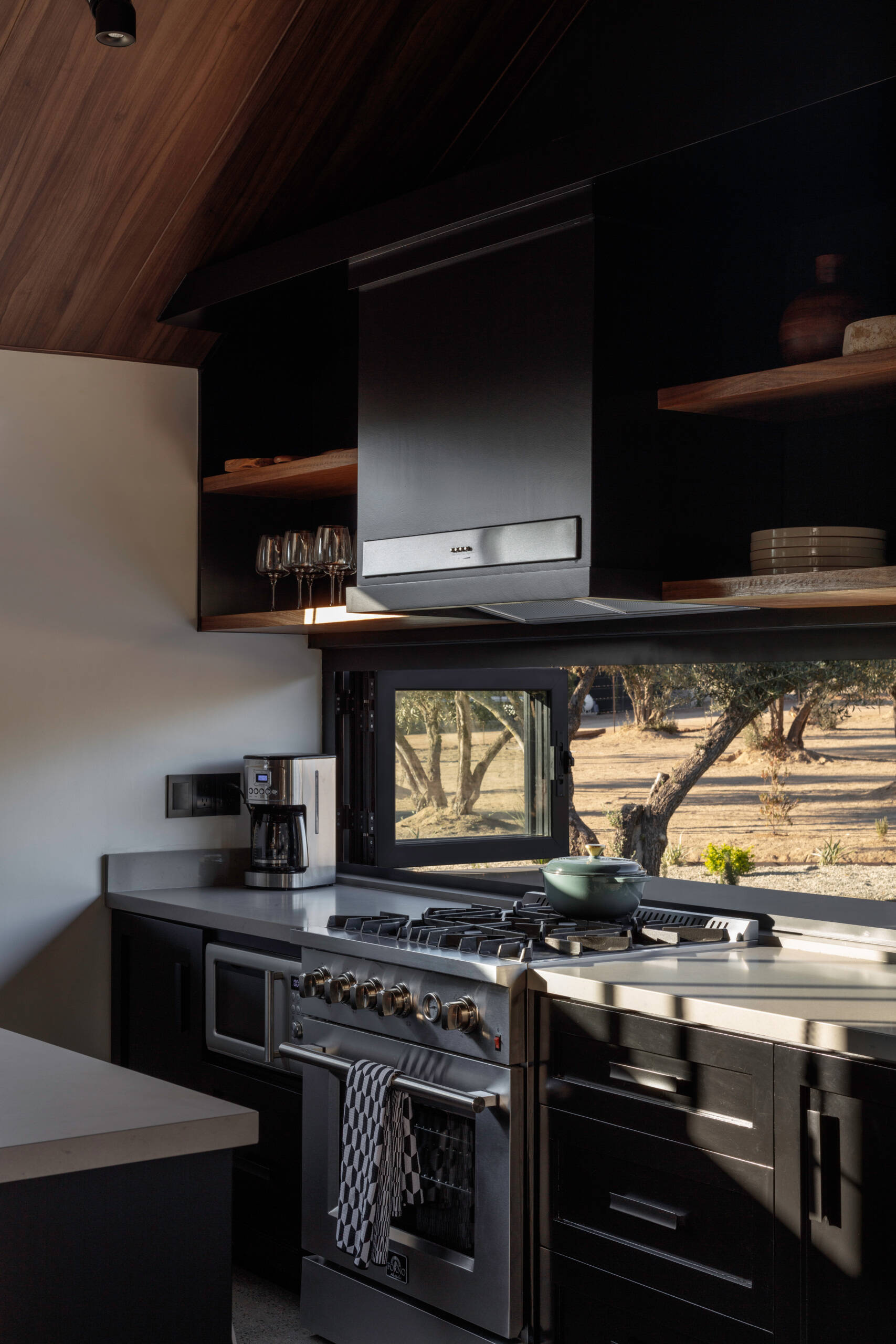Explore the modern black kitchen at Serentha Cabin by ORTO STUDIO in Baja California, Mexico. Matte black cabinets, a large gray kitchen island, leather-woven bar stools, glass pendant lights, stainless steel appliances, and open timber shelves create a stylish cabin kitchen with views of the olive grove.