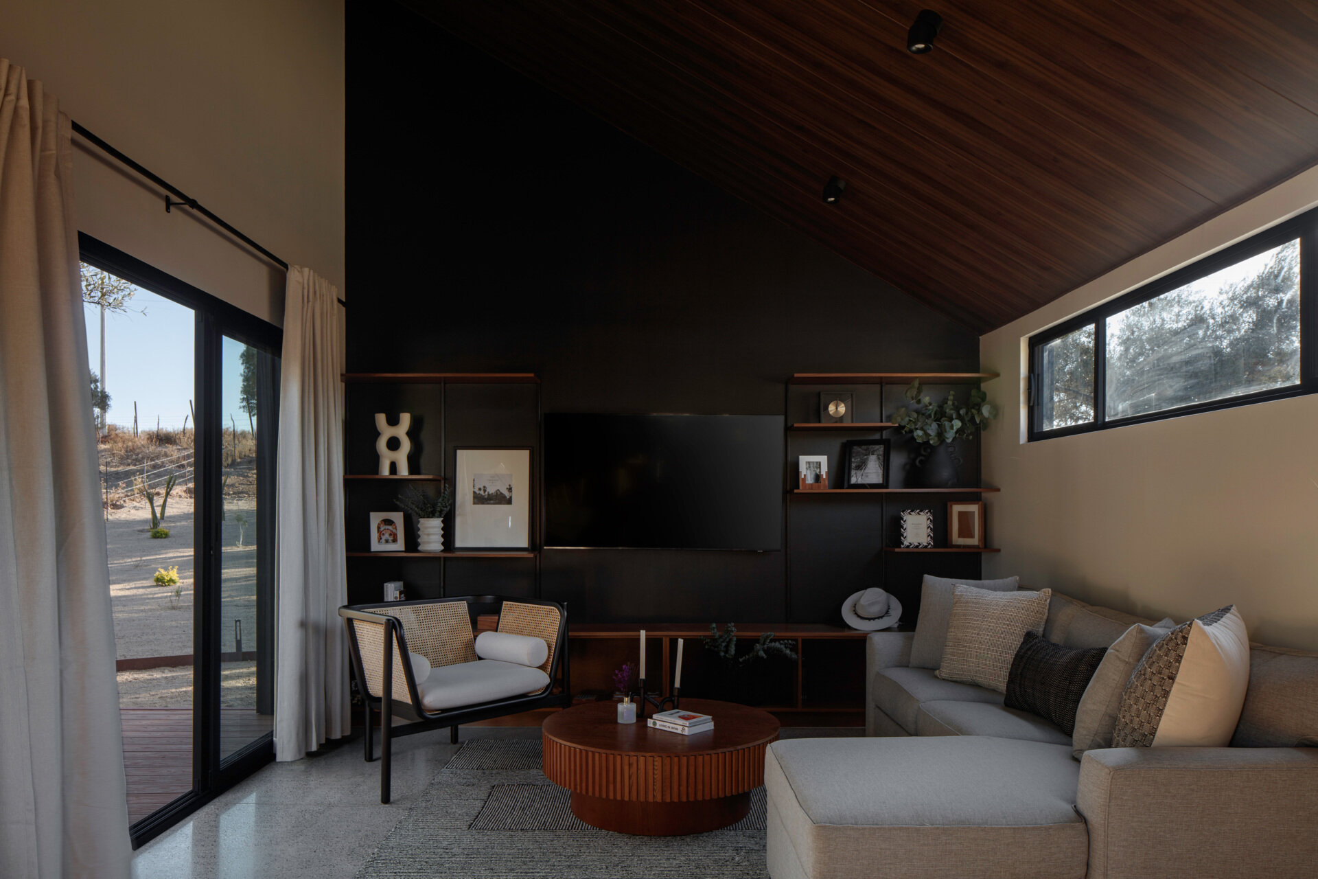 A sloping timber ceiling, polished concrete floors, dark built-in shelving, a gray sectional sofa, and warm wood furniture create a welcoming living room.
