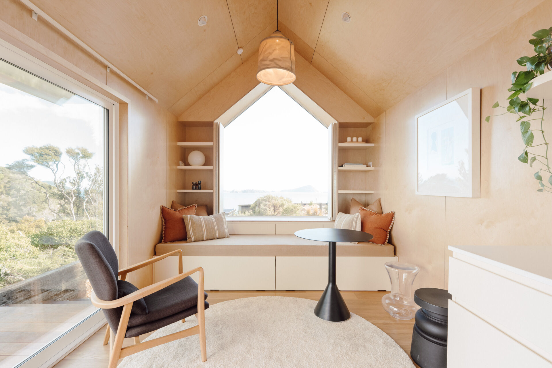 Inside Arc's small coastal cabin, a beautiful birch plywood living room features a built-in window bench, hidden guest sleeping space, open shelving and panoramic harbour views. Inspiration for small living rooms and window seat designs.