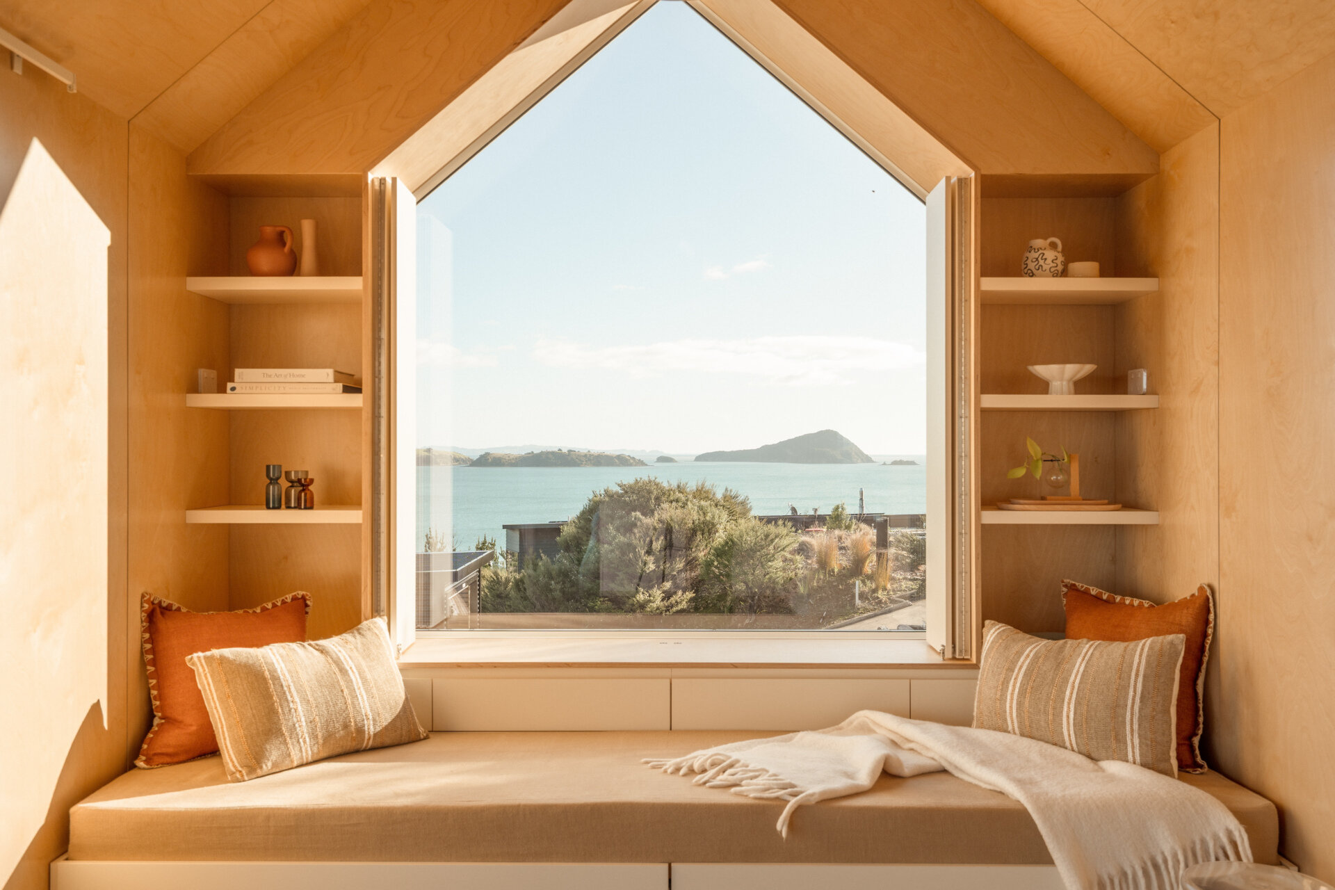 Inside Arc's small coastal cabin, a beautiful birch plywood living room features a built-in window bench, hidden guest sleeping space, open shelving and panoramic harbour views. Inspiration for small living rooms and window seat designs.