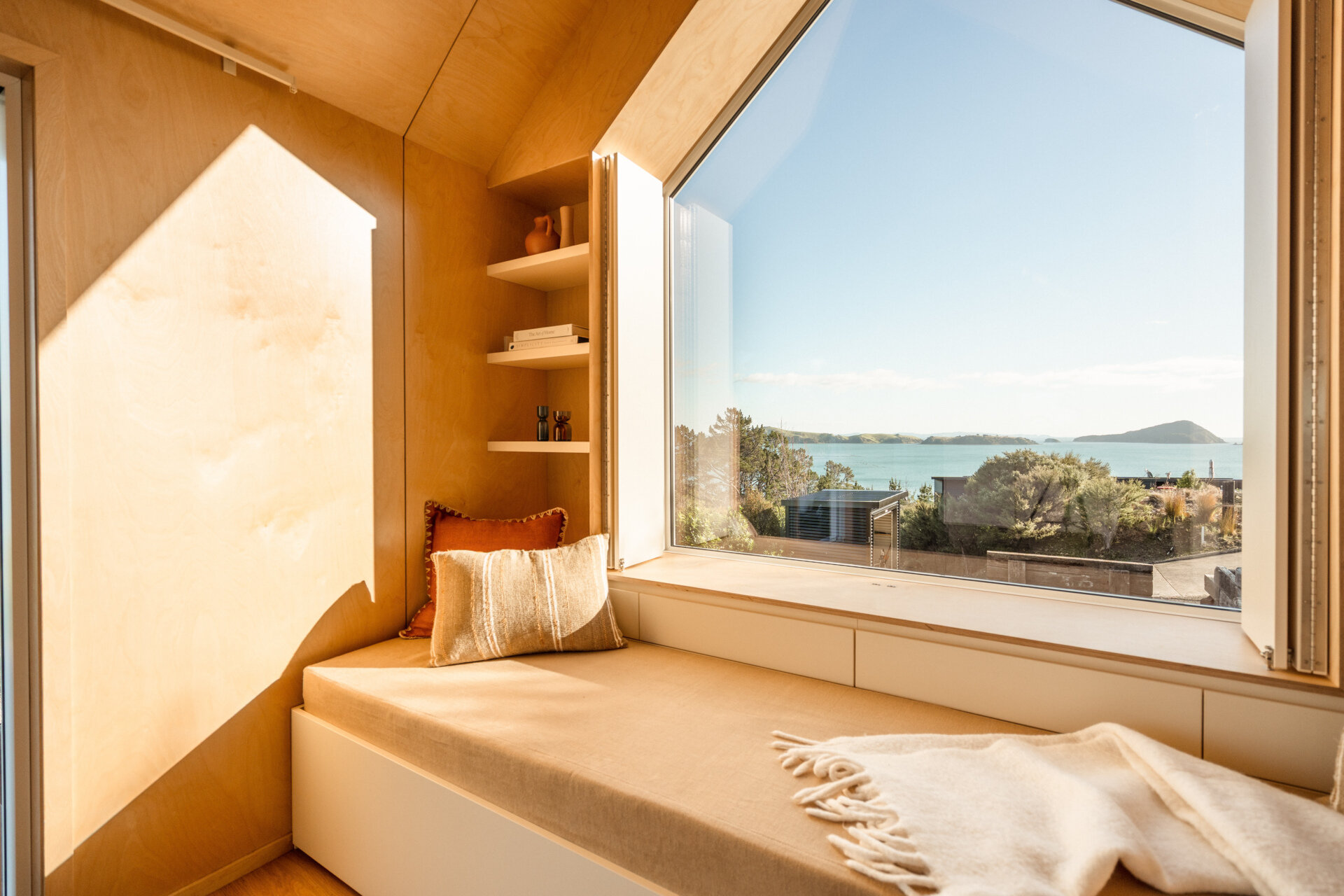 Inside Arc's small coastal cabin, a beautiful birch plywood living room features a built-in window bench, hidden guest sleeping space, open shelving and panoramic harbour views. Inspiration for small living rooms and window seat designs.