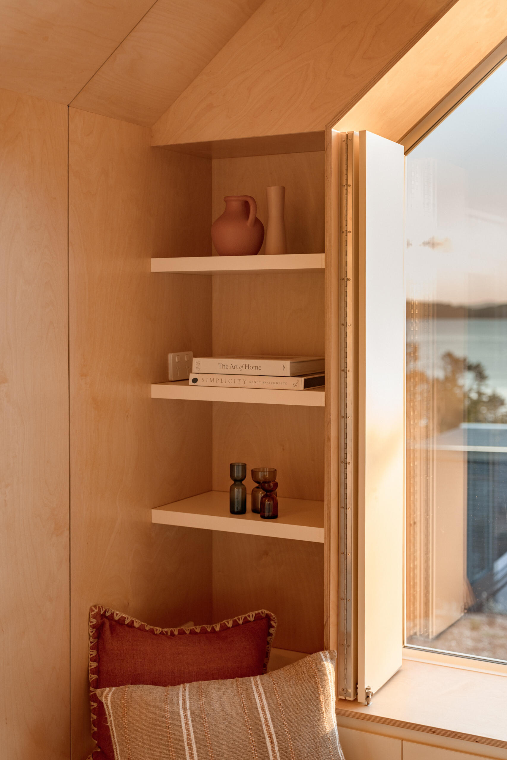 Inside Arc's small coastal cabin, a beautiful birch plywood living room features a built-in window bench, hidden guest sleeping space, open shelving and panoramic harbour views. Inspiration for small living rooms and window seat designs.
