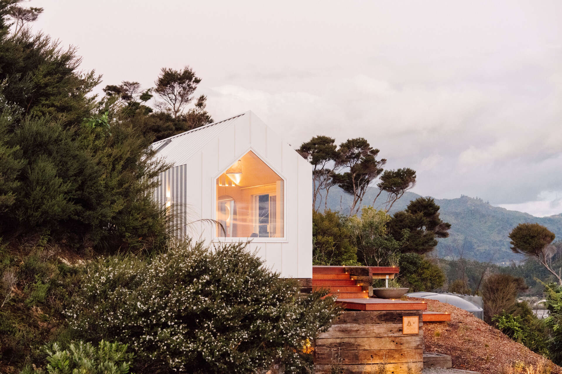 Discover the architecture of Arc's Wyuna Bay cabin in New Zealand, featuring white standing-seam metal cladding, a gabled roof, expansive glazing and timber decking overlooking Coromandel Harbour.