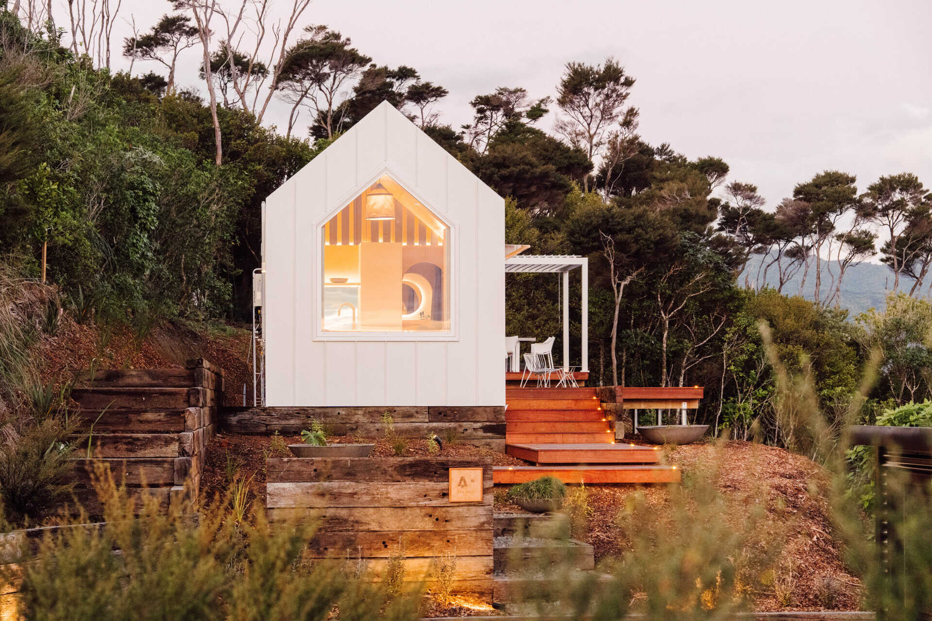 Discover the architecture of Arc's Wyuna Bay cabin in New Zealand, featuring white standing-seam metal cladding, a gabled roof, expansive glazing and timber decking overlooking Coromandel Harbour.
