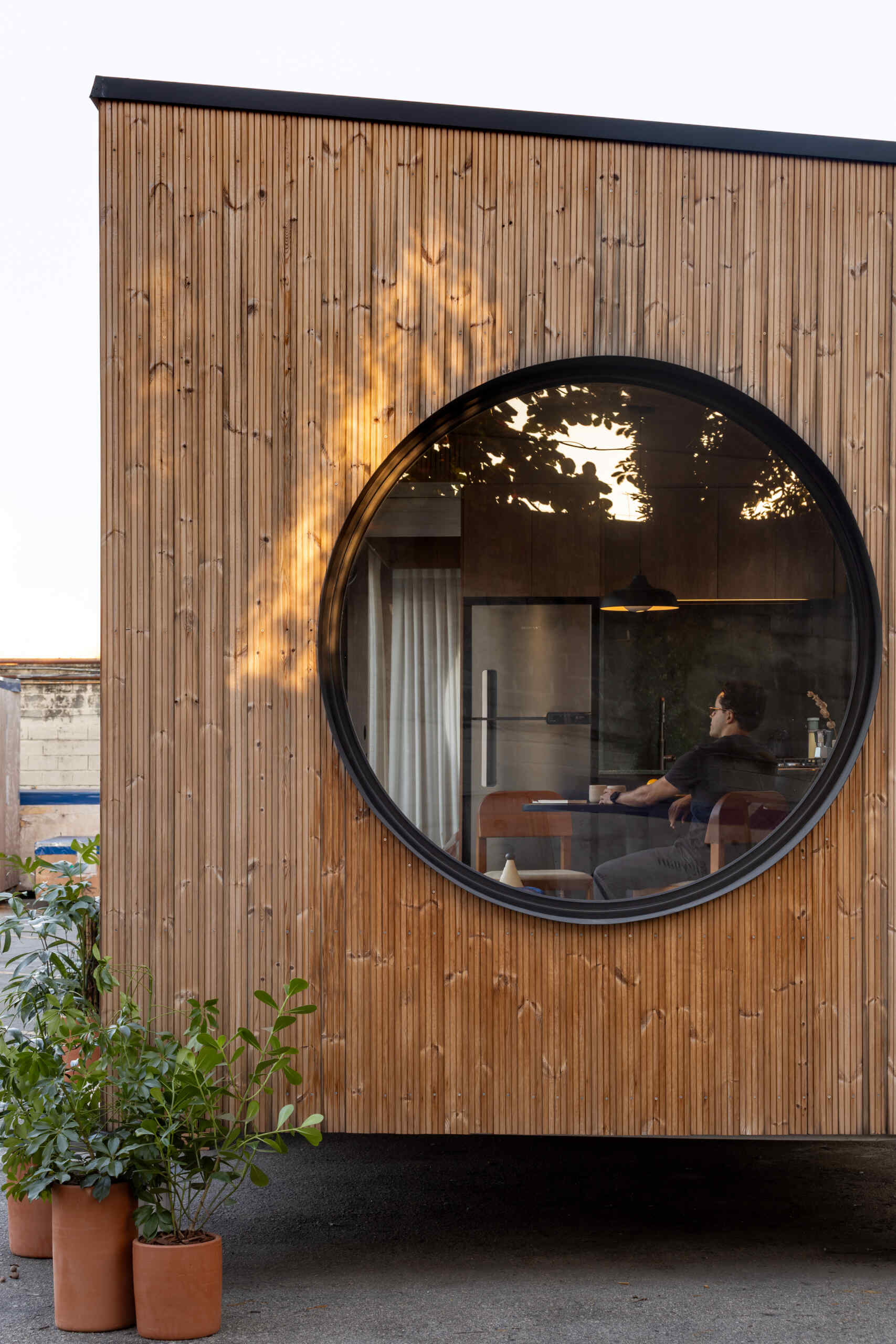 This small prefabricated house features vertical wood cladding, large sliding glass doors, and distinctive circular windows at both ends.