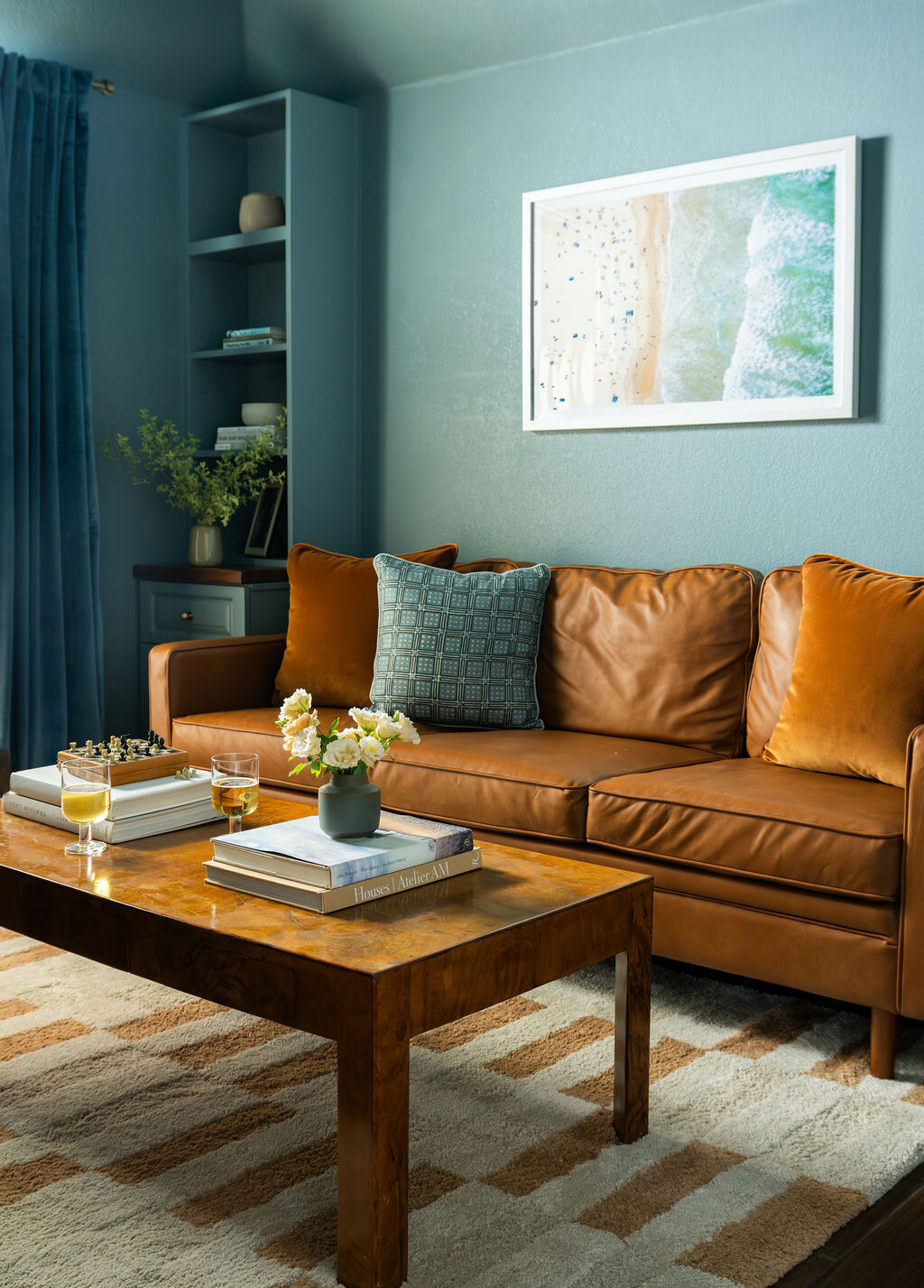 A cozy sitting area within a blue-green California home office, featuring a brown leather sofa, velvet cushions, and a wood coffee table. Built-in shelving, framed artwork, and a patterned cream rug complete the family lounge.