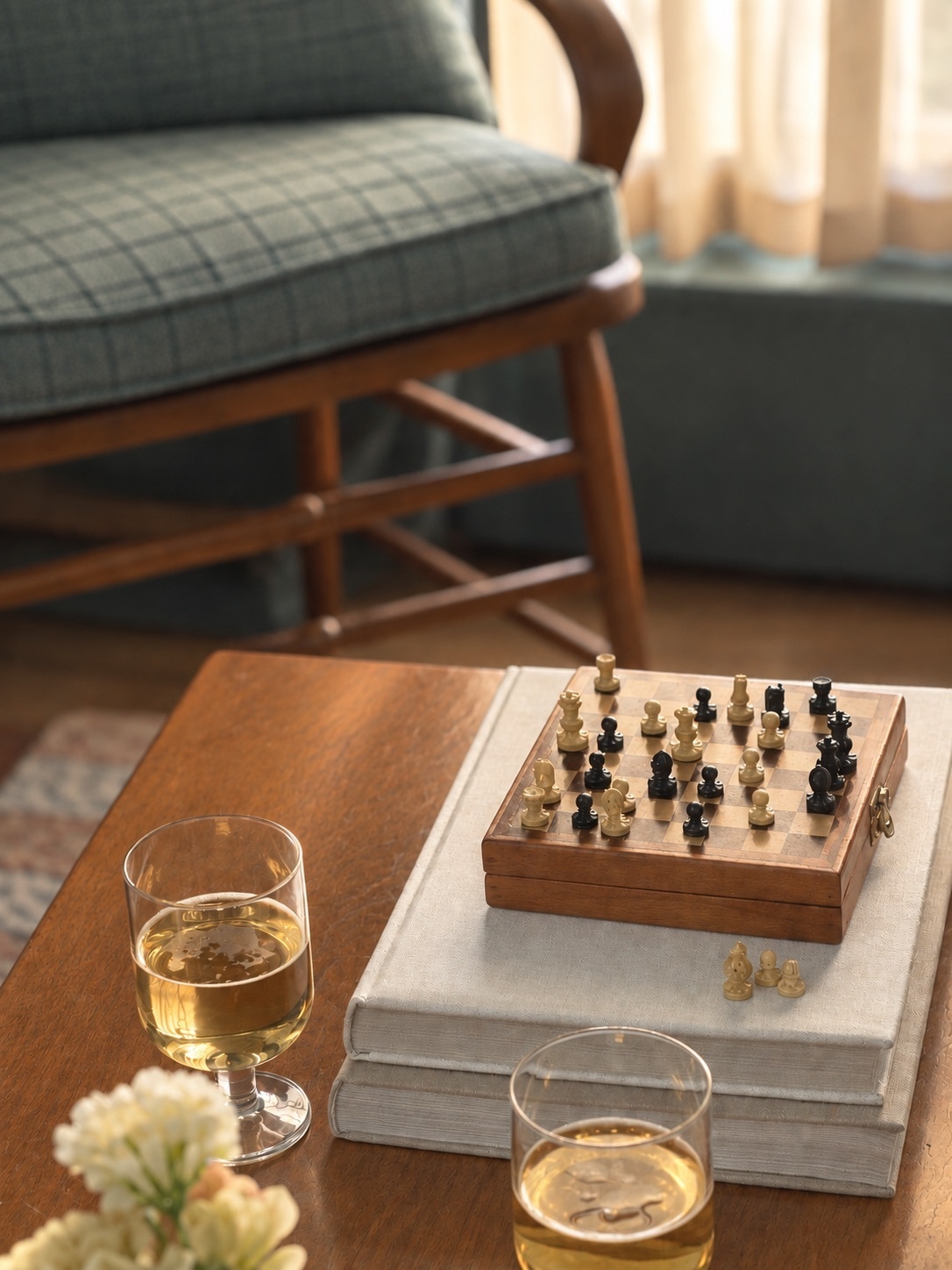A close-up of a wood coffee table in a family lounge, styled with a small chess set, stacked books, and drinking glasses. A vintage wood chair with a checked cushion appears in the background, adding to the room's traditional character.
