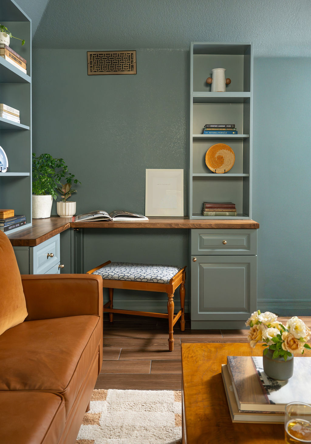 Custom home office cabinetry combines blue-green bookshelves, traditional cabinet fronts, and warm wood desktops arranged around a corner. An upholstered wood stool fits beneath the desk, while a brown leather sofa and coffee table sit nearby.