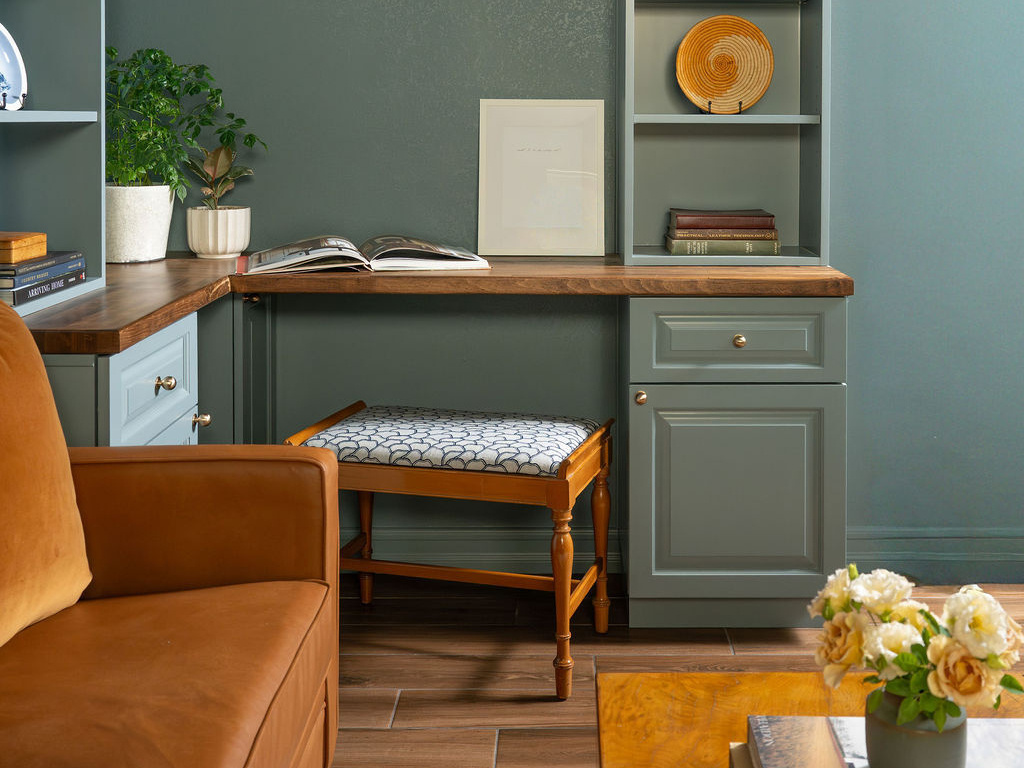 A small home office in California featuring blue-green built-in cabinetry, wood desktops, open bookshelves, and a vintage upholstered stool. The color continues across the walls and ceiling, creating a coordinated workspace.