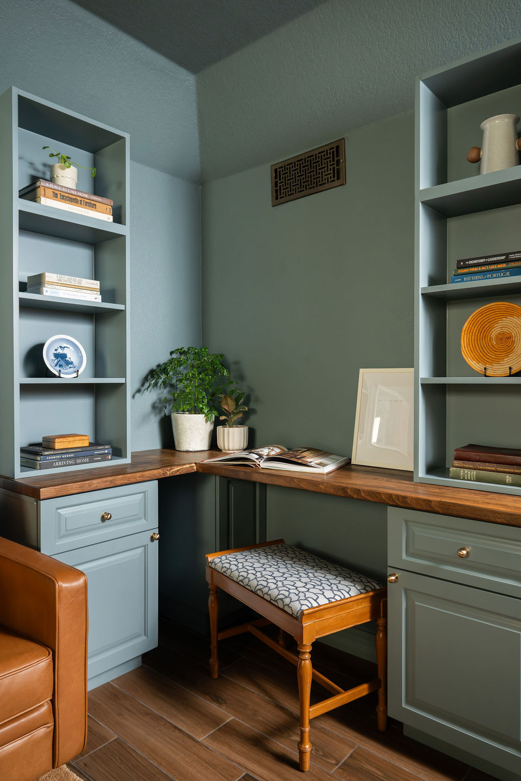 A compact L-shaped home office desk surrounded by tall blue-green bookshelves and custom storage cabinets. Wood work surfaces, brass cabinet knobs, and a vintage upholstered stool add warmth to the colorwashed interior.