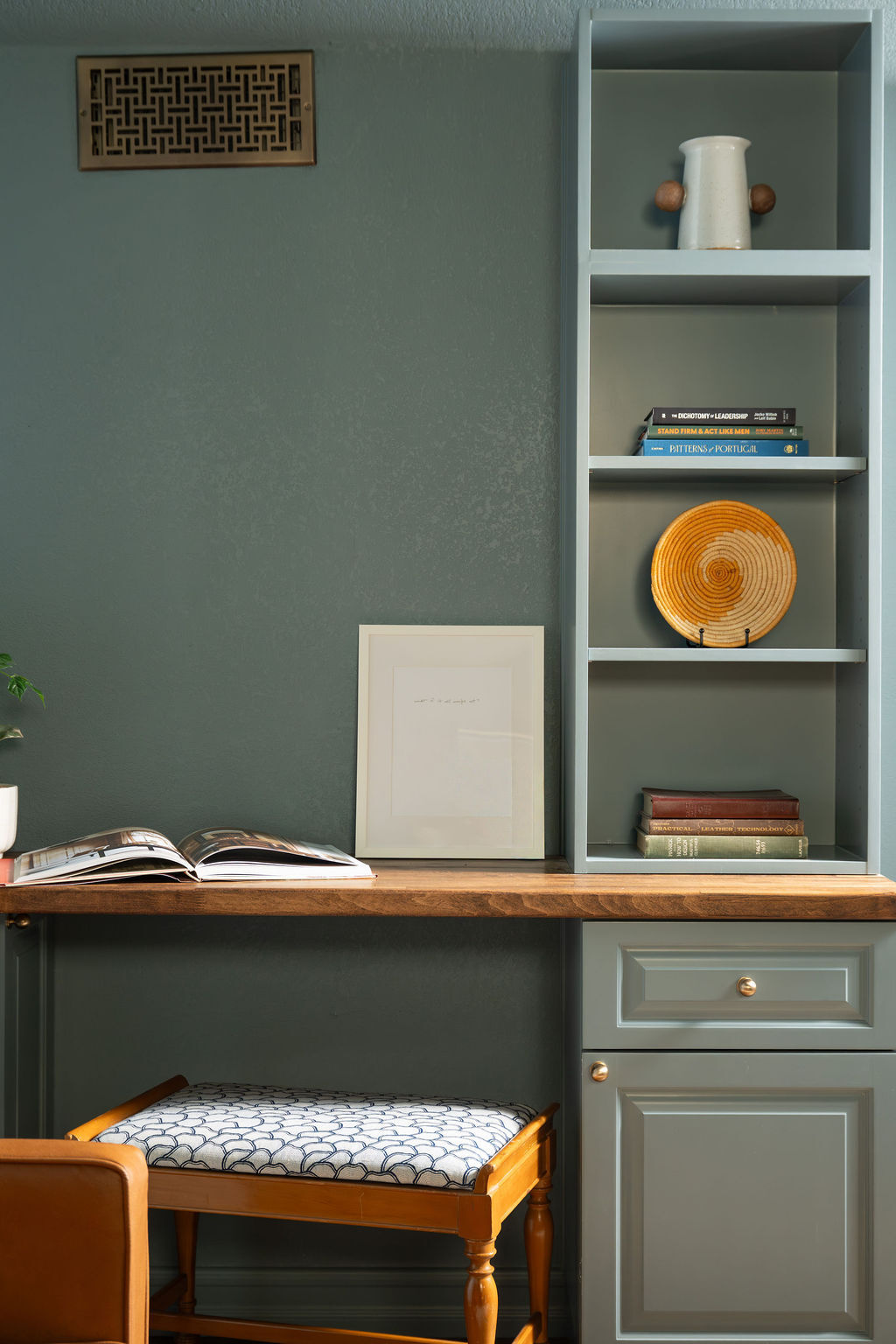 A built-in home office desk with blue-green cabinetry, open display shelves, a wood desktop, and an upholstered stool. Books, pottery, and an orange decorative plate add small accents of color to the matching walls and shelving.