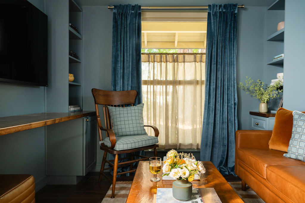 A small multipurpose home office with blue-green built-in cabinetry, a wall-mounted television, and a brown leather sofa. Blue curtains frame the window, while a wood armchair and coffee table create a seating area for family movie nights.