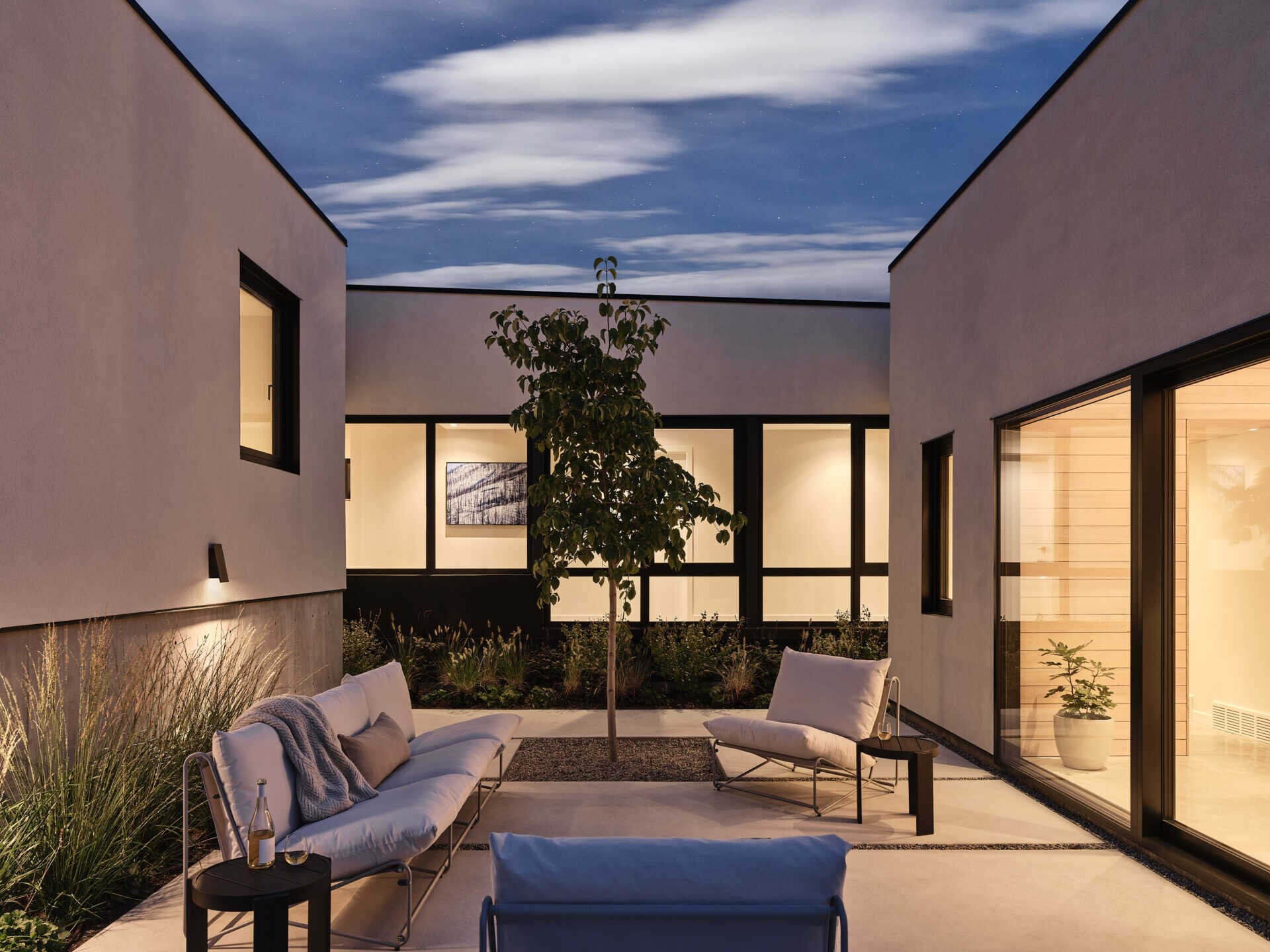A modern courtyard lounge surrounded by white stucco walls and large glass doors, photographed at dusk. White outdoor sofas, lounge chairs, a small tree, and planted borders create a sheltered seating area between the house's separate wings.