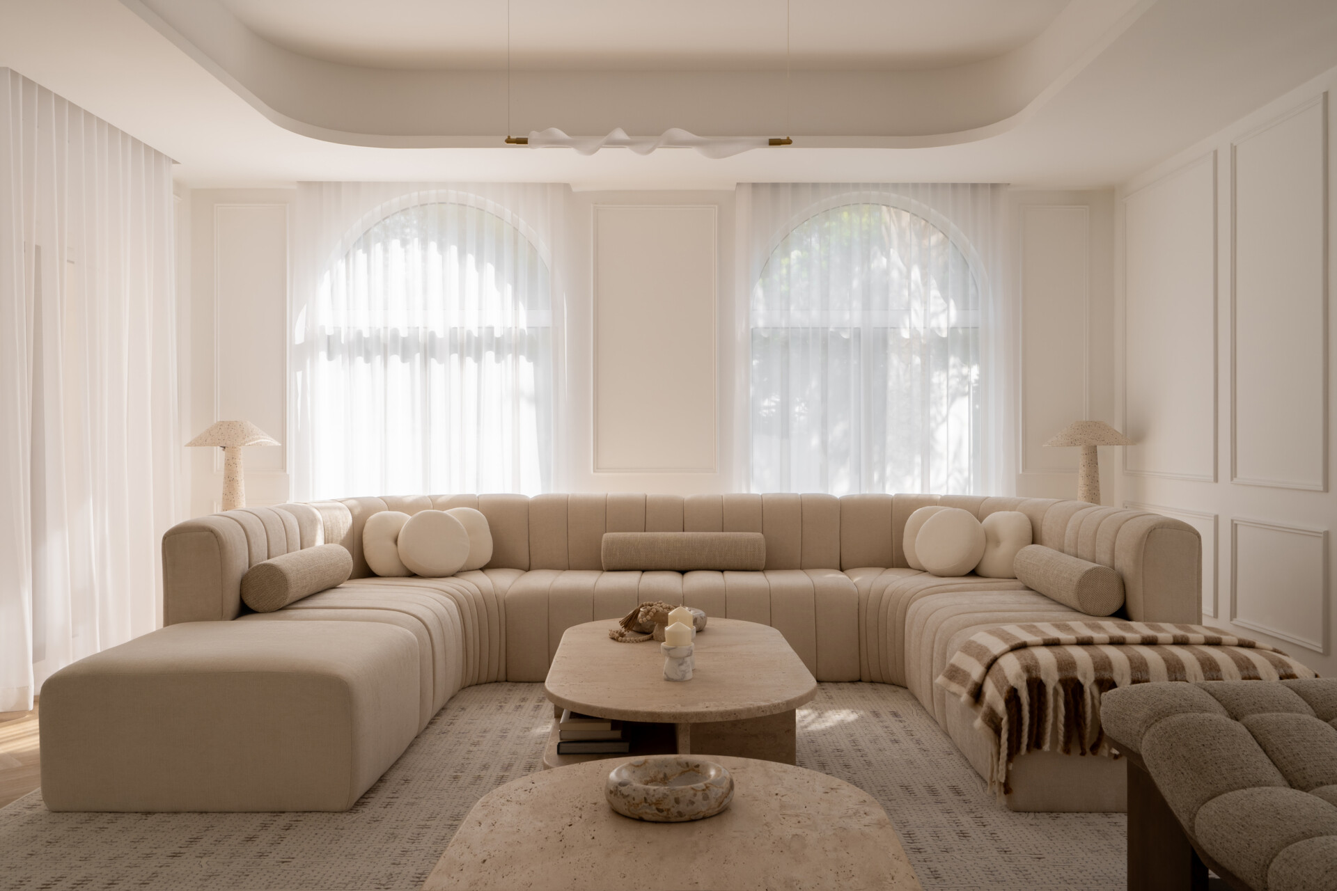 Step inside the media room of The White Arcade in Dubai. A large curved sectional sofa, bouclé armchairs, stone coffee tables, textured rug, and arched windows create a cosy yet sophisticated neutral living room designed by D’Ora Tokai Designs.