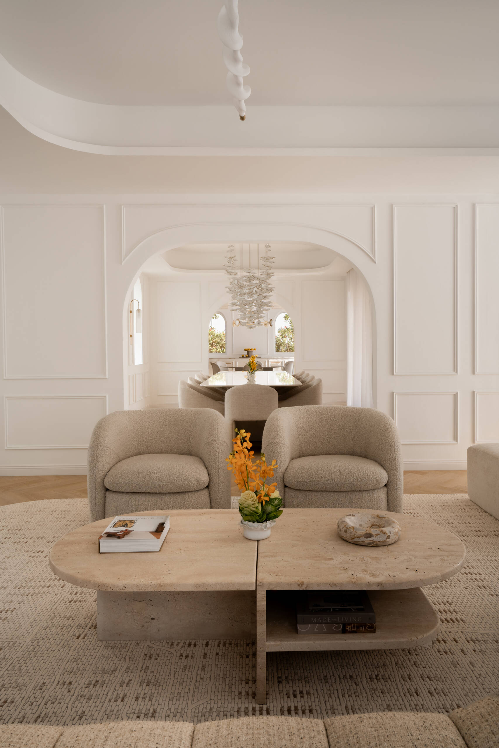 Step inside the media room of The White Arcade in Dubai. A large curved sectional sofa, bouclé armchairs, stone coffee tables, textured rug, and arched windows create a cosy yet sophisticated neutral living room designed by D’Ora Tokai Designs.