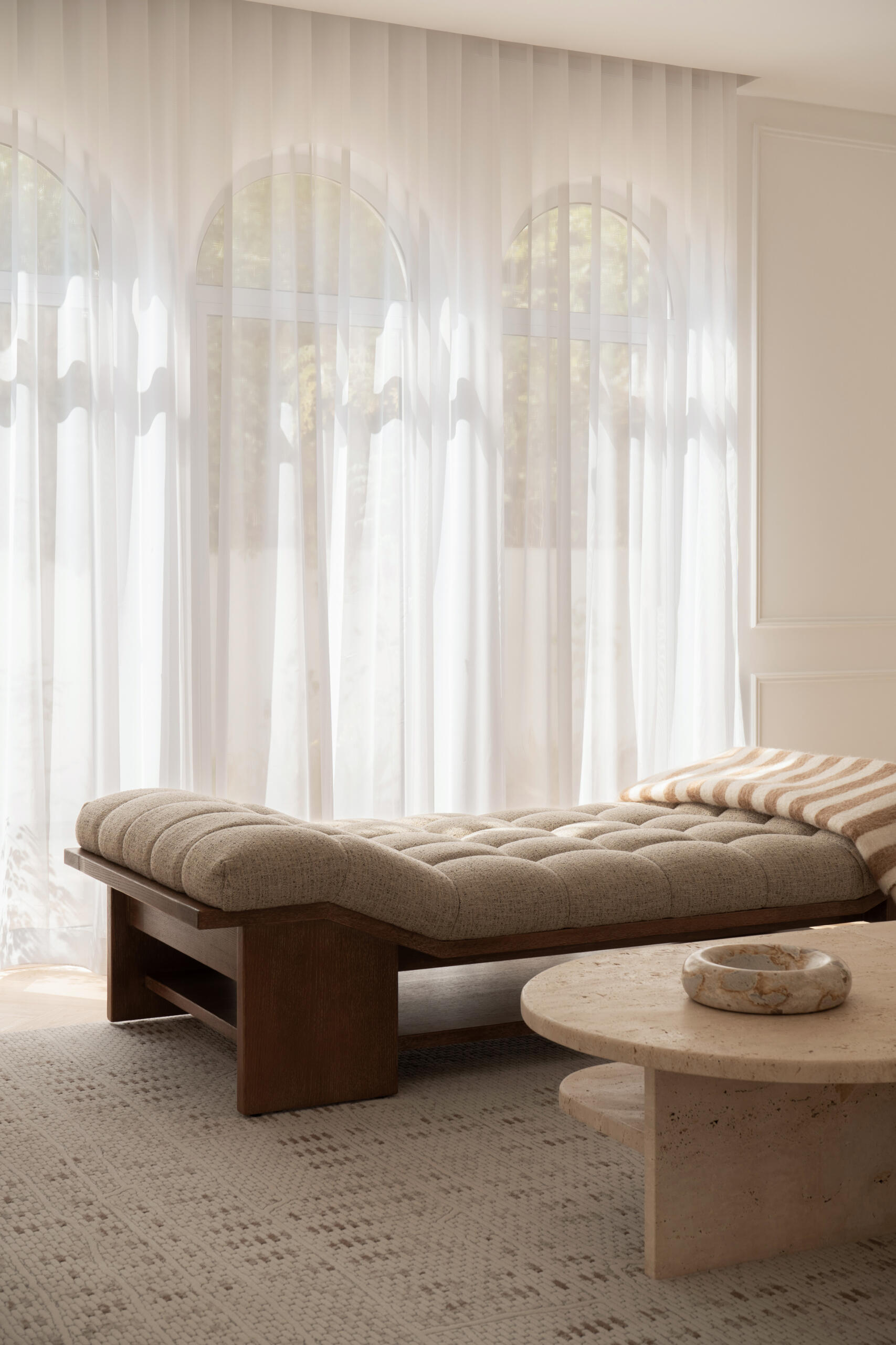 Step inside the media room of The White Arcade in Dubai. A large curved sectional sofa, bouclé armchairs, stone coffee tables, textured rug, and arched windows create a cosy yet sophisticated neutral living room designed by D’Ora Tokai Designs.