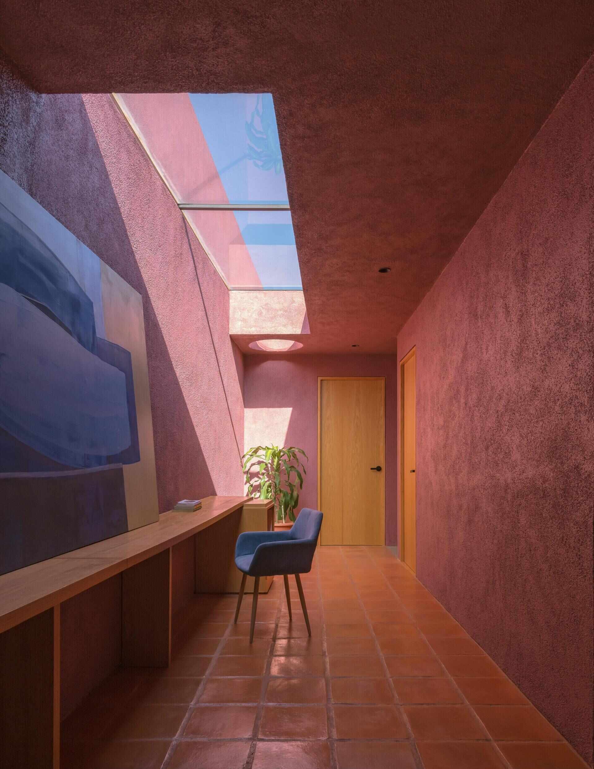 A mezzanine home office in a modern Mexican house features a long built-in wood desk, overhead skylight, pink walls, blue artwork, and a small sitting area overlooking the living room.