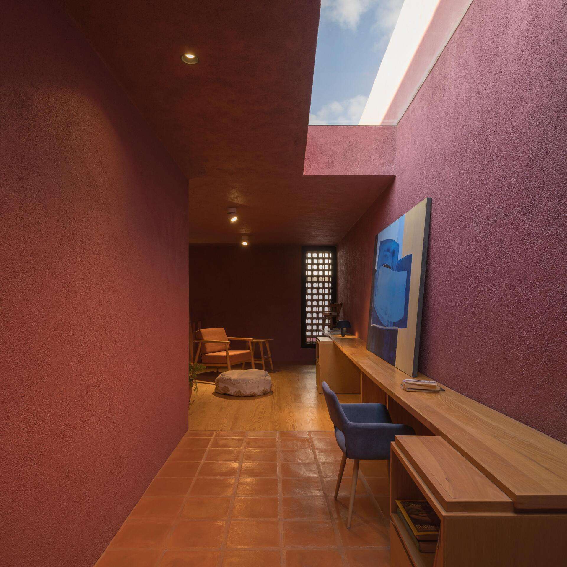 A mezzanine home office in a modern Mexican house features a long built-in wood desk, overhead skylight, pink walls, blue artwork, and a small sitting area overlooking the living room.