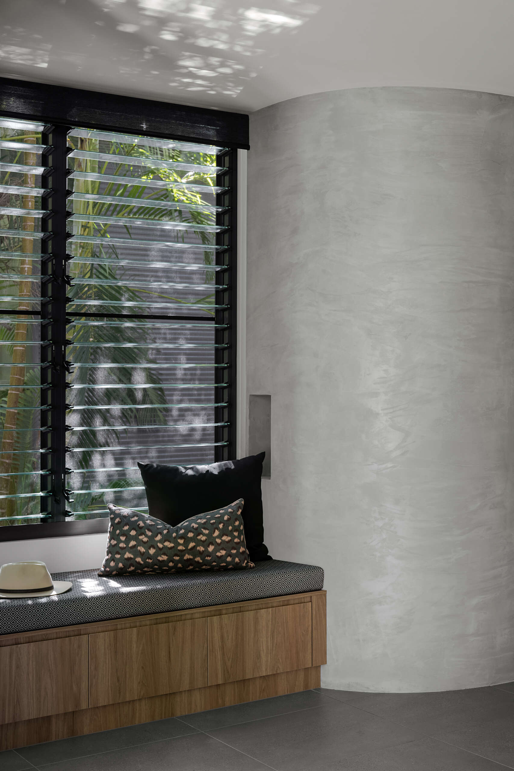 Discover the dining area of Raintree in Noosa Heads, Australia, featuring a natural timber dining table, black upholstered chairs, a modern circular pendant light, botanical artwork, and a built-in window seat overlooking tropical greenery.